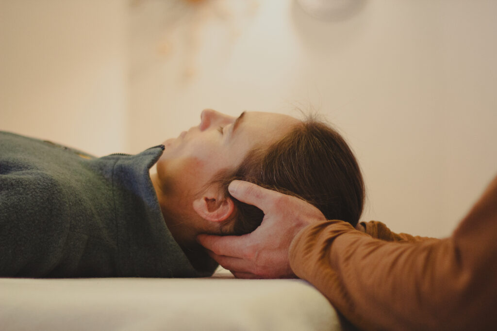 Eine Frau liegt während einer Craniosacralen Behandlung entspannt auf einer Liege, der Therapeut hält ihren Schädel.